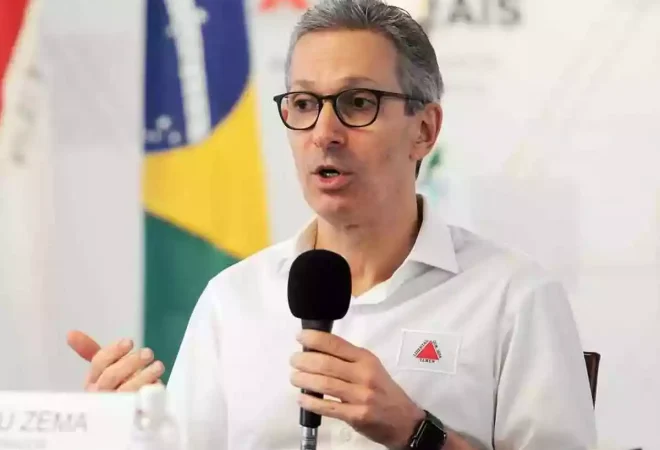 romeu-zema-sentado-em-uma-mesa-com-um-microfone-na-mao-e-as-bandeiras-de-minas-gerais-e-do-brasil-ao-fundo_1_52937