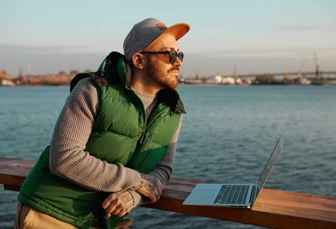 In search of inspiration. Outdoor picture of handsome fashionable guy with neat beard and tattooed hands standing by city river, working distantly on portable electronic gadget, using free WiFi