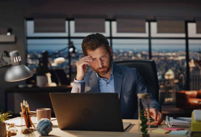 Serious boss checking phone sms in late office interior closeup. Focused business man typing laptop keyboard. Busy manager pondering project working at panoramic windows evening workplace alone