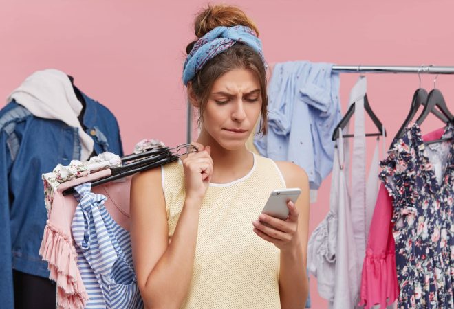 Serious female model standing agianst lay figure and rack of clothes, holding hangers with clothes in one hand and smart phone reading about discounts in clothes shop. People, shopping, technology