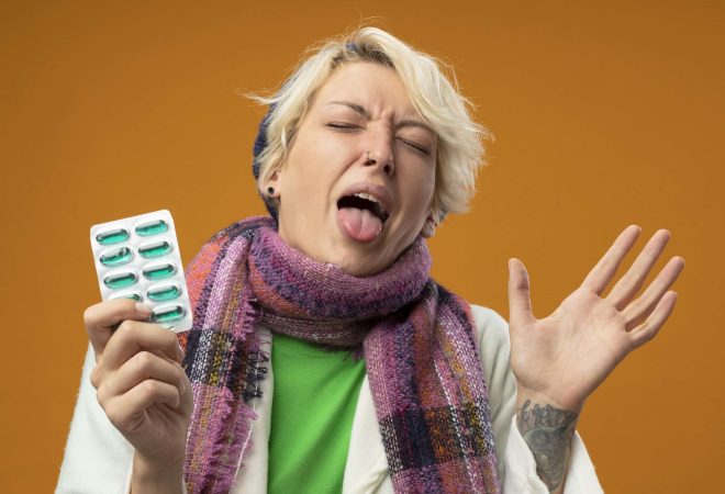 sick unhealthy woman with short hair in warm scarf and hat holding blister with pills raising arm sticking out tongue with disgusted expression standing over orange background