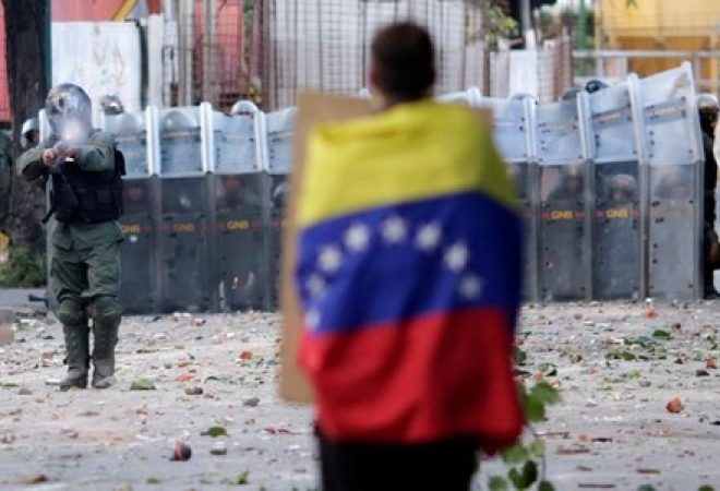 Homem com bandeira da Venezuela parado em frente a forças de segurança contra protestos durante manifestação contra o governo do presidente Nicolas Maduro em Caracas, na Venezuela