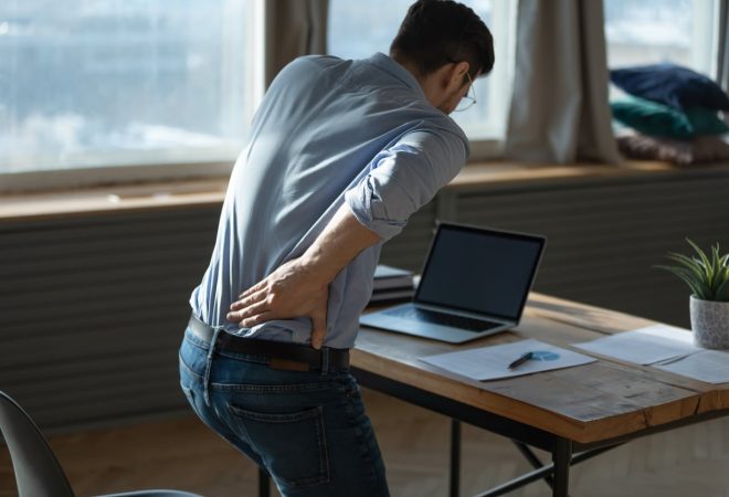 Rear view young man suffering from sudden backache, getting out of uncomfortable chair at workplace, touching lower back, unhealthy businessman student office worker feeling discomfort