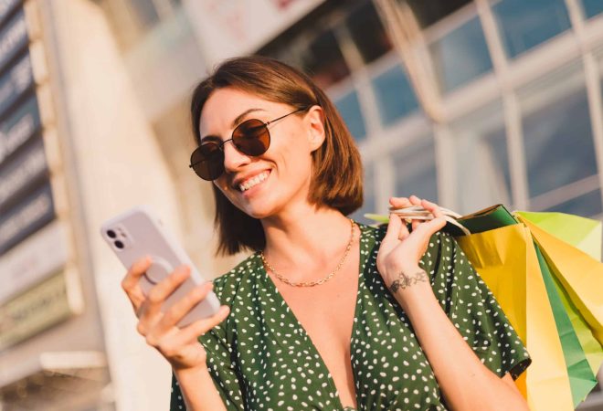 Woman at sunset with colorful shopping bags and parking lot by shopping mall happy with mobile phone