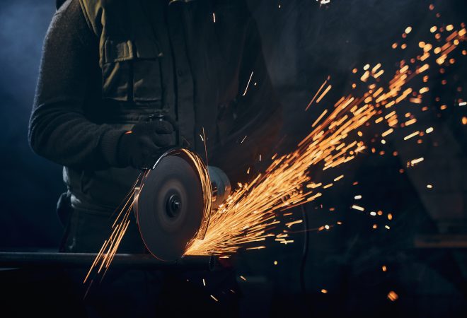 Close up of man in special dark suit and black protective gloves working angle grinder for metal with large flash sparks. Concept of process working electric steel cutter machine.