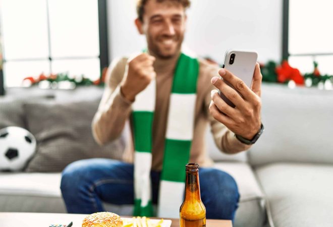 Young hispanic man supporting soccer match using smartphone sitting by christmas decor at home