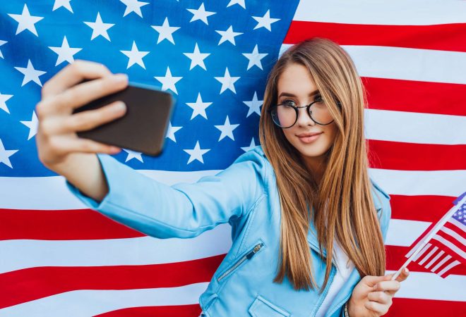 Tsu is the US Independence Day! Young pretty American girl doing selfie against the background of the flag.